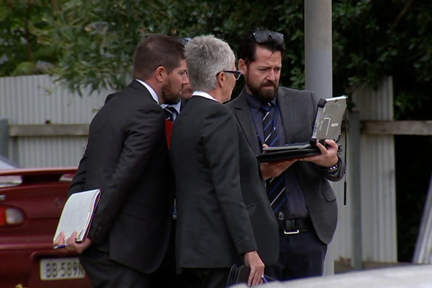 A group of detectives wearing suits huddle around an ipad at the scene of an assault