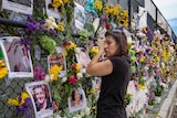 A woman wipes tears from her face as she looks along a chainlink fence adorned with flowers and photos arranged as a vigil.