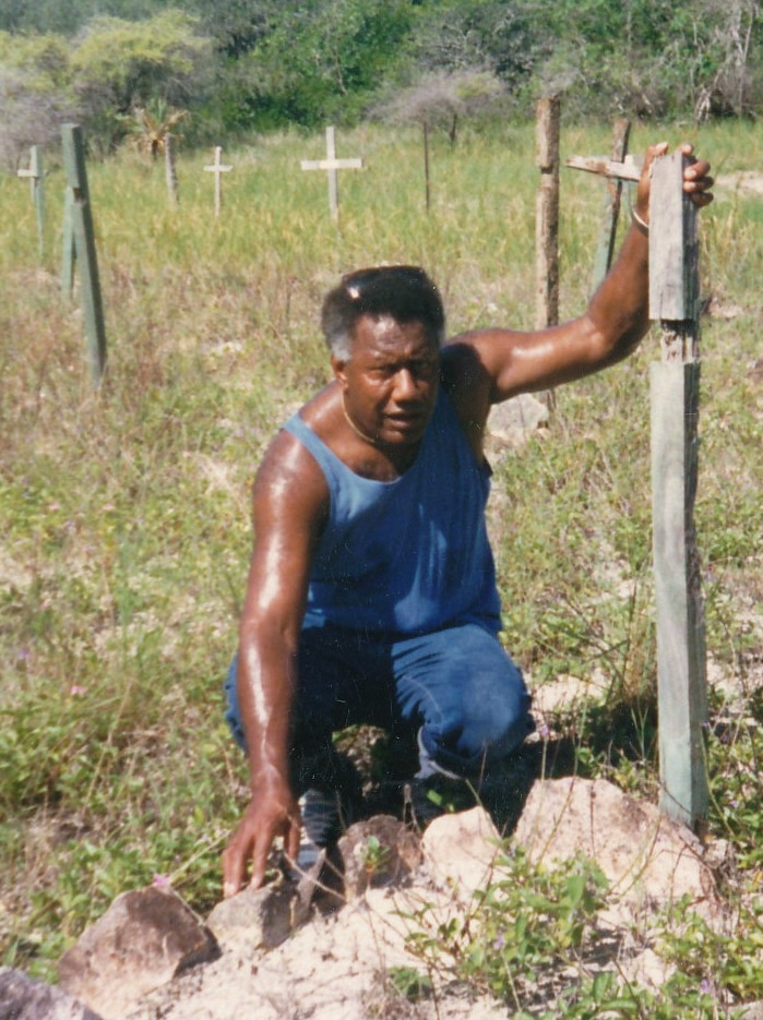 In a photo from 1988, an indigenous man wearing a blue singlet kneels at a gravesite in a cemetery overgrown with grass.