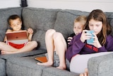 Three young children sit on a couch engrossed in phones and tablets.