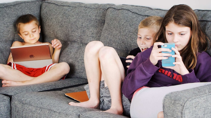 Three young children sit on a couch engrossed in phones and tablets.