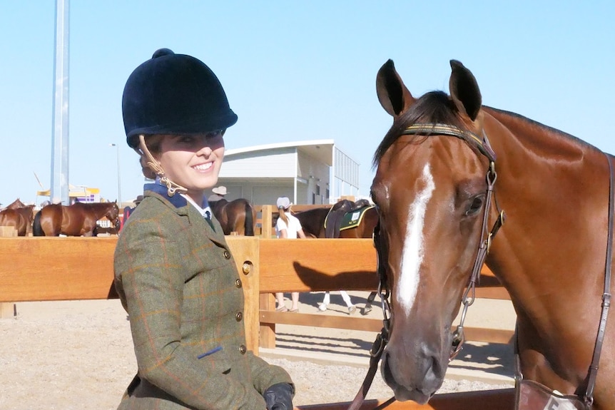 April Docherty stands against a wooden rail beside her bay horse.