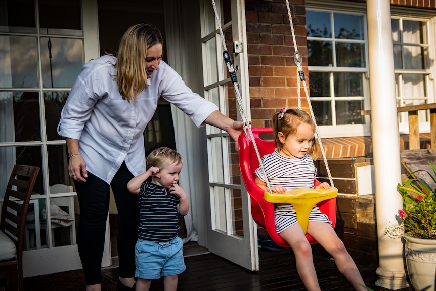 Robyn pushes Emilia on a bright rd swing, while little Mathew watches curiously.