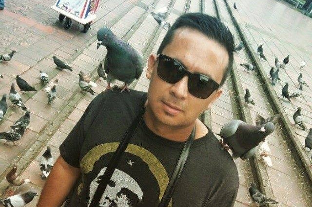 A man stands in a city square surrounded by pigeons, with a pigeon on each shoulder.