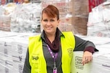 Kate has red hair tied up and she's wearing a fluro yellow vest and is leaning on boxes on food. She is smiling.