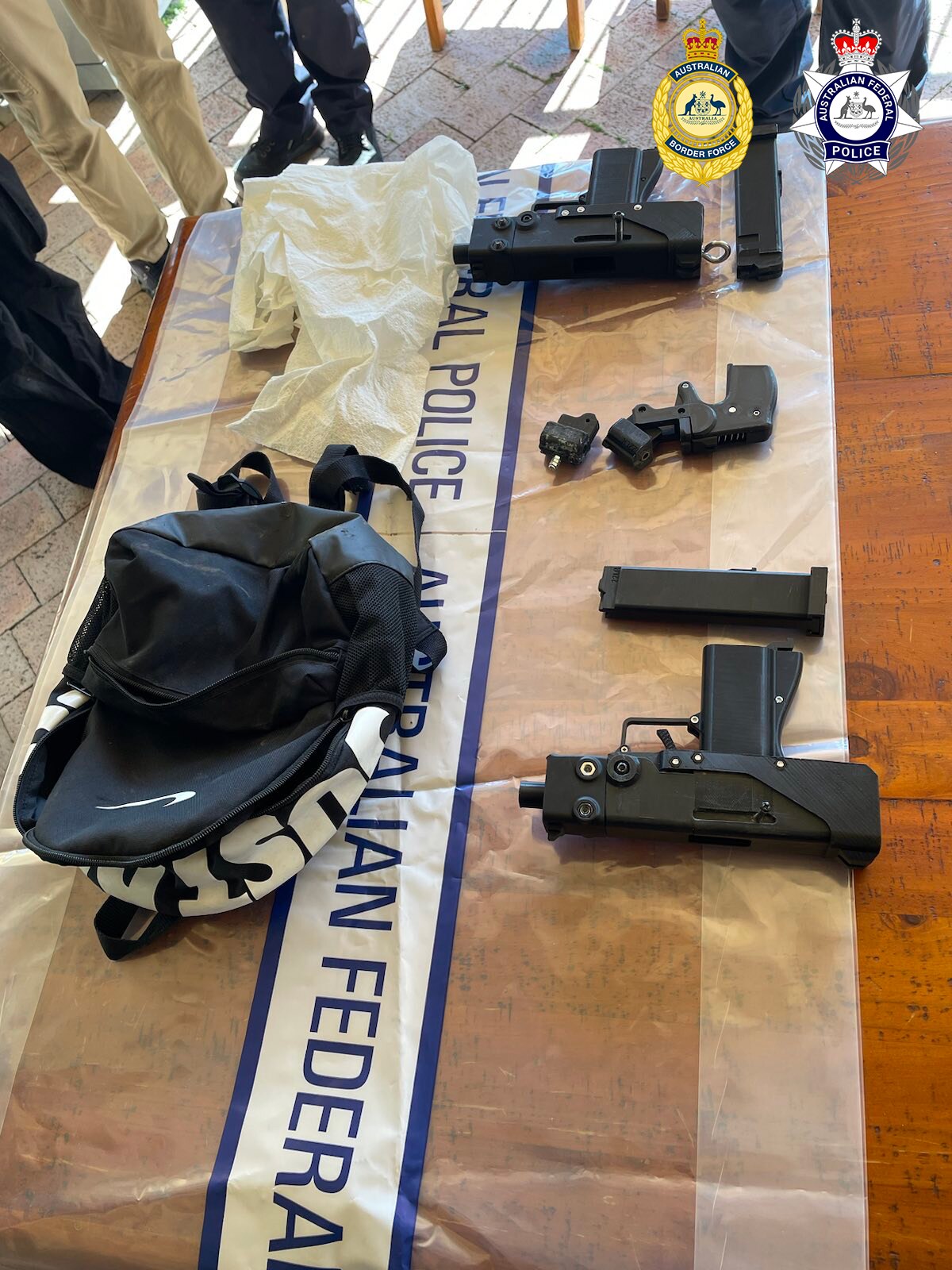 Multiple black guns on the floor next to a black backpack. 