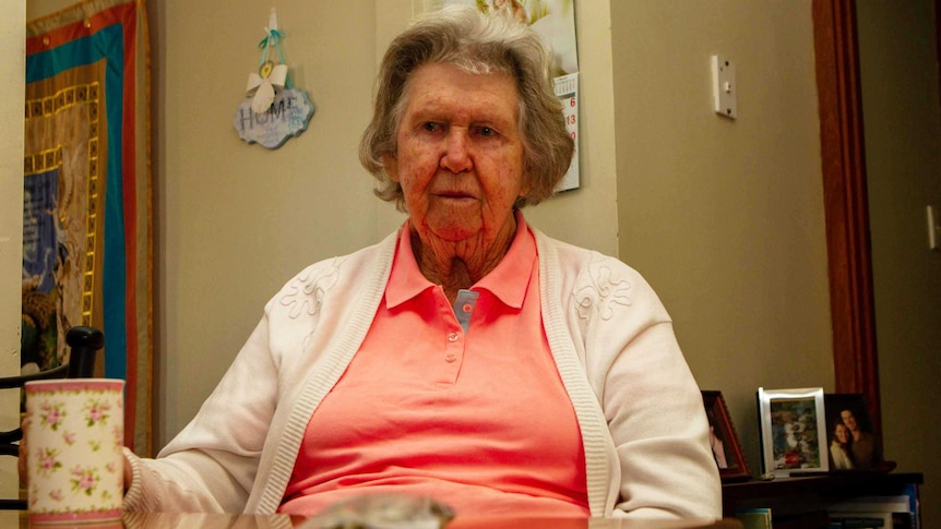 An elderly woman sits at a kitchen table. An undistinguishable bag sits on the table in front of her.