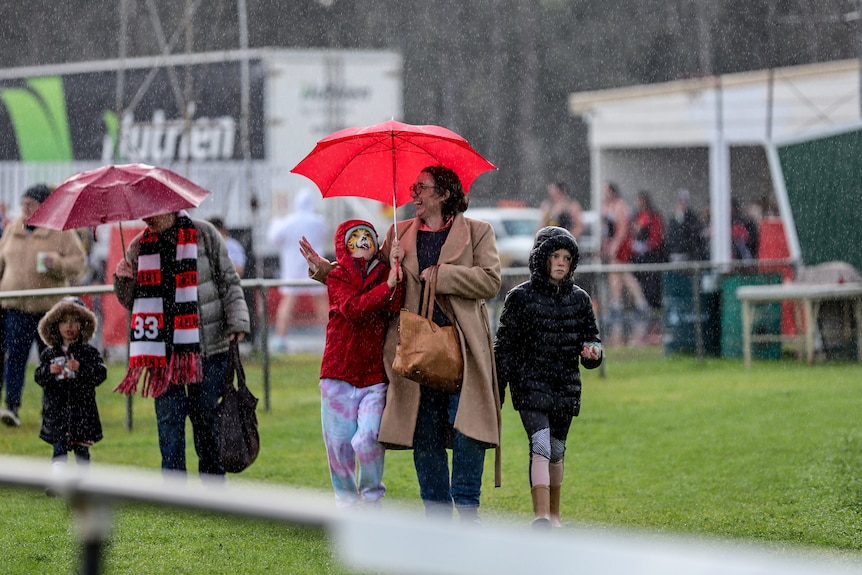 Woman in coat holds red umbrella with two children next to her walking across football ground. 