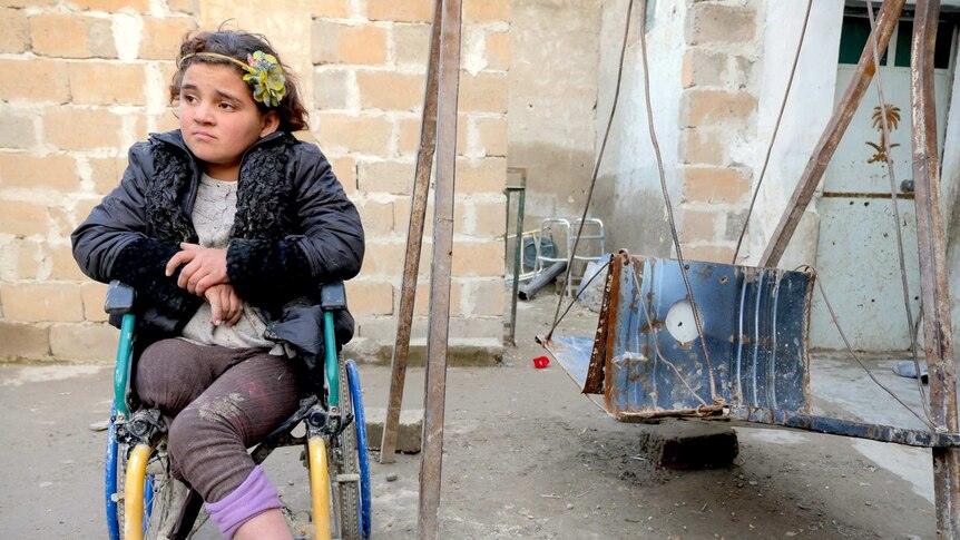 A girl missing a leg sits in a wheelchair in a rubble-filled courtyard
