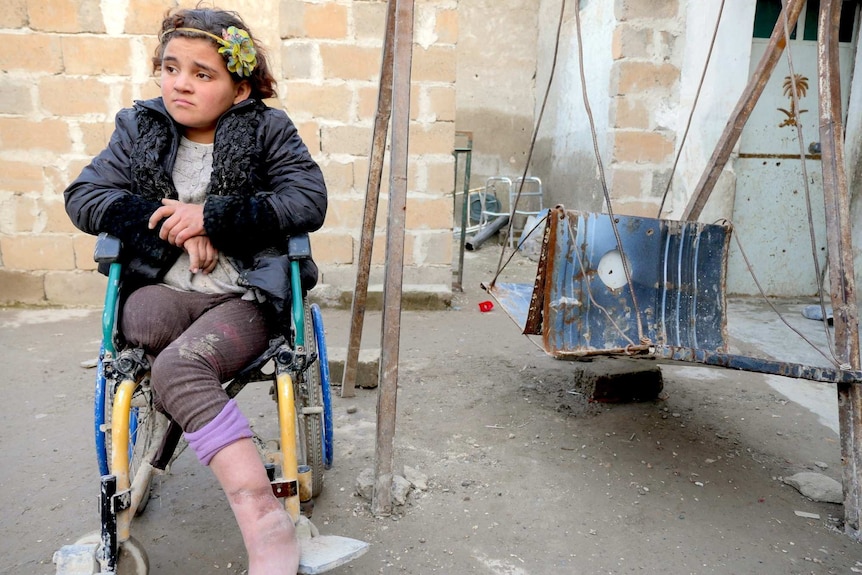 A girl missing a leg sits in a wheelchair in a rubble-filled courtyard