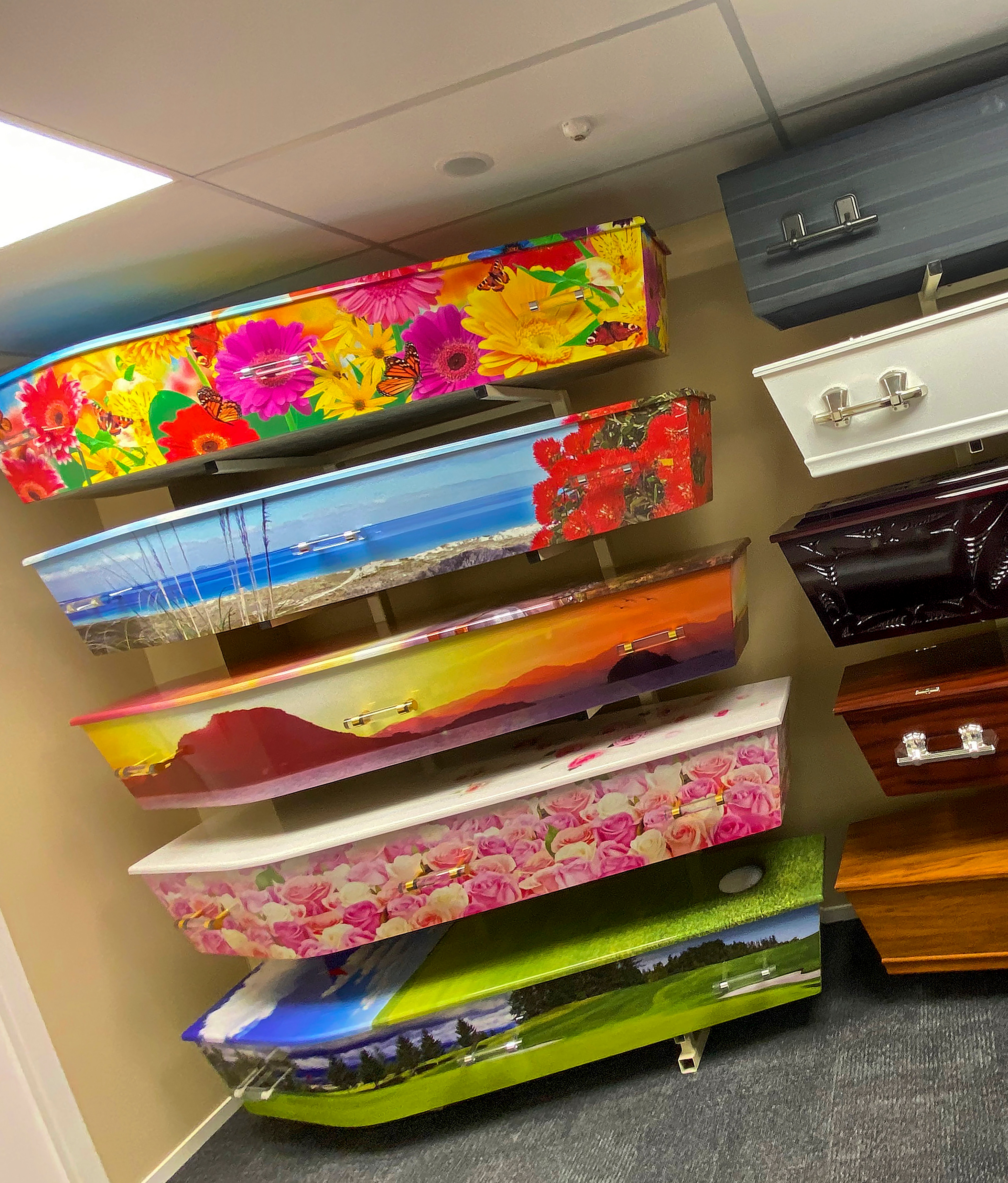 A range of colourful coffins are mounted on a wall. Some are floral, others with a beach scene