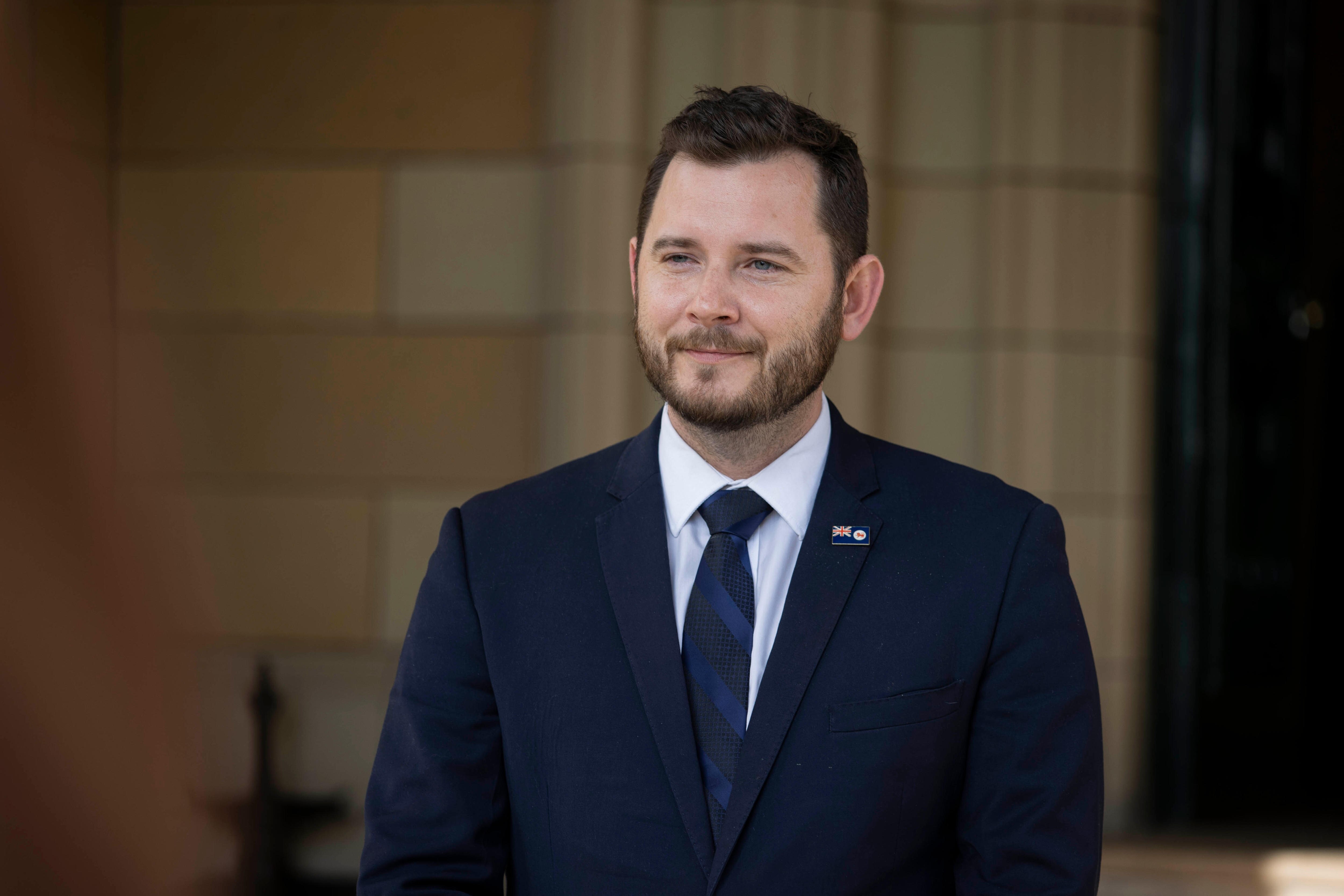 A man with a cropped beard wearing a suit stands in a sandstone portico