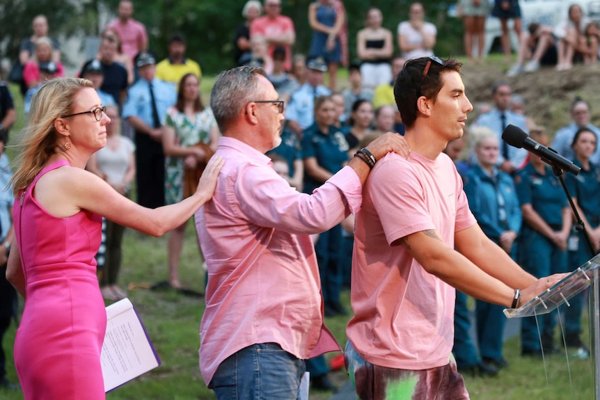 Three people dressed in pink lean on each other as one speaks into a microphone