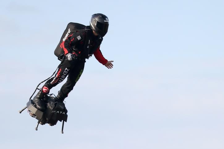 Franky Zapata is seen on the left hand side of the frame in mid air. He wears a black helmet and black and red full body suit.