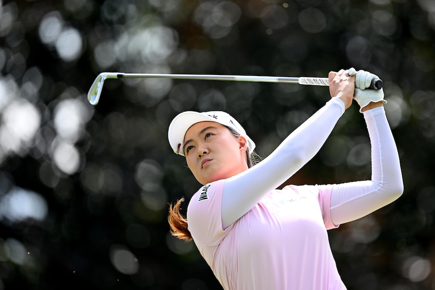 Woman in pink shirt and cap swings a golf club, looks out to distance 