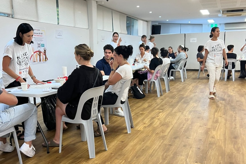 People sitting at trestle tables in a large hall getting the inside of their cheeks swabbed