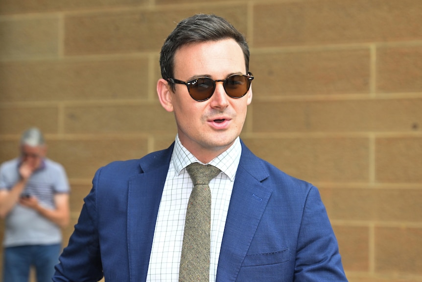 Sam wears a suit and tie and sunglasses and walks along mid-speech. He has short brown hair.