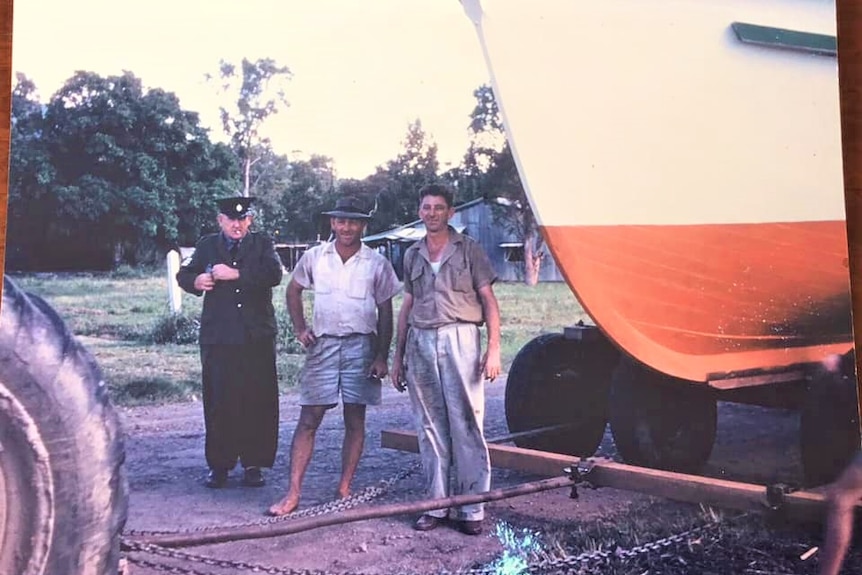An old photo of two men and a policeman in uniform stand next to a very large orange and white boat keel, on a huge trailer.
