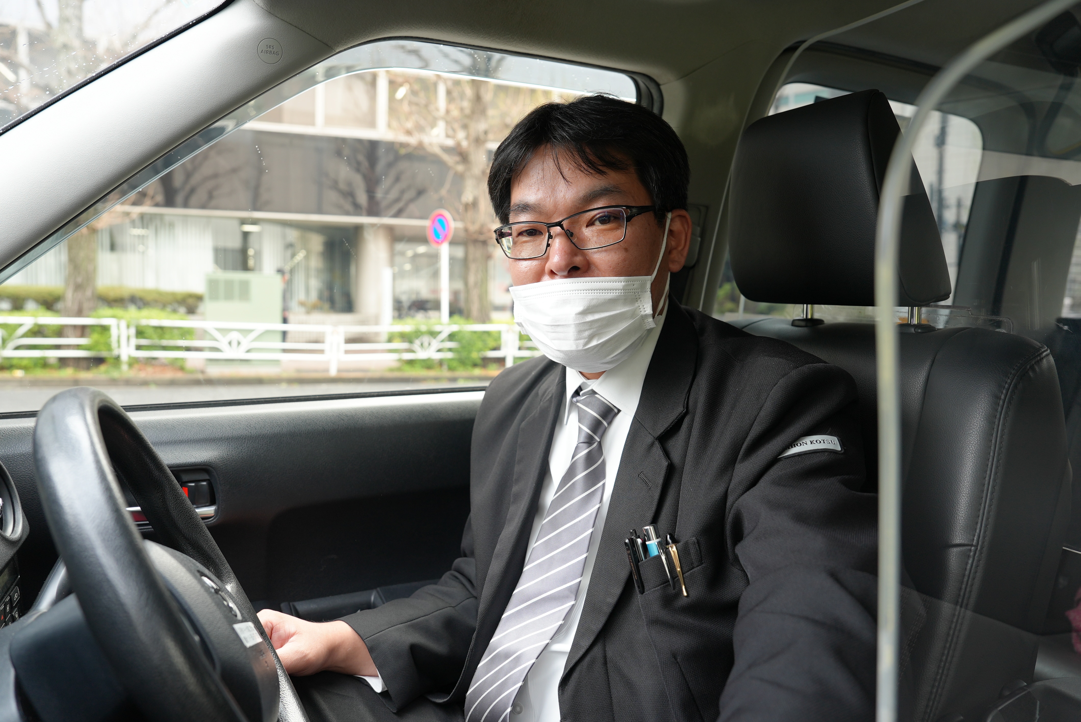 A japanese man wearing a mask sits in in the drivers seat of a taxi.