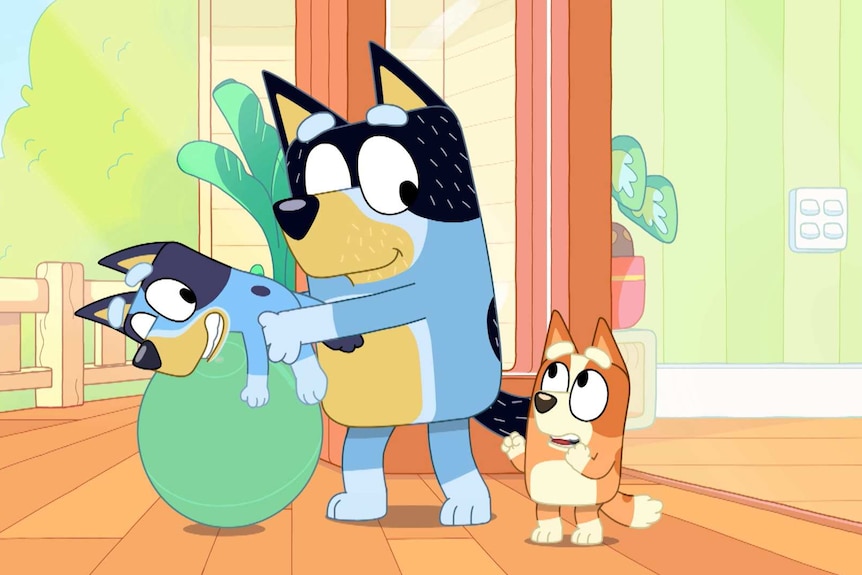 A scene from Bluey in which Bandit bounces Bluey on a yoga ball while Bingo looks on.