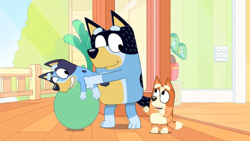 A scene from Bluey in which Bandit bounces Bluey on a yoga ball while Bingo looks on.