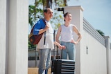 A man and a woman stand at the front gate of a house holding luggage and looking into the distance.