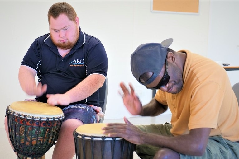 Two men sitting on chairs playing large African drums inside an open room.