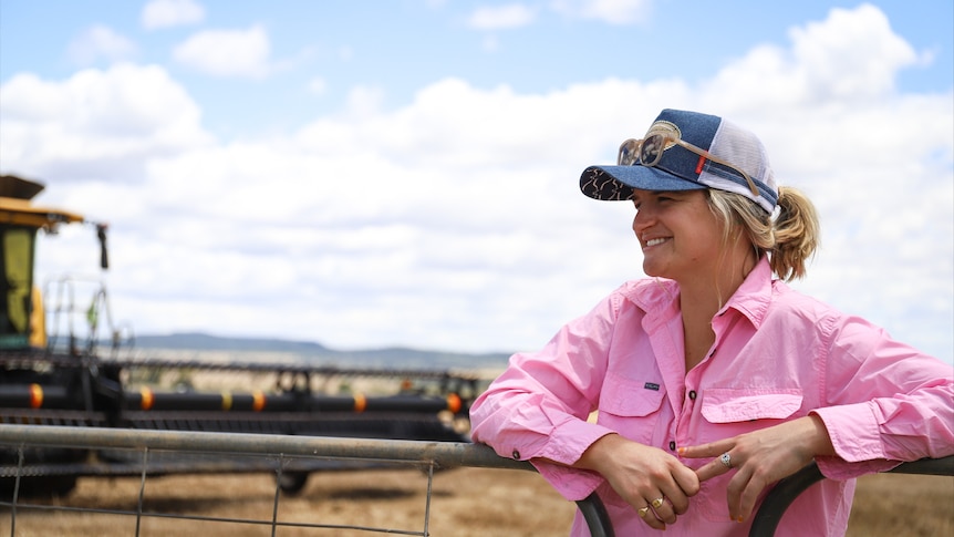 A woman wearing a cap and a pink shirt stands on a farm with her hands on a railing.