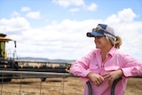 A woman wearing a cap and a pink shirt stands on a farm with her hands on a railing.