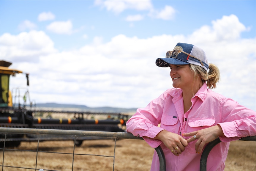A woman wearing a cap and a pink shirt stands on a farm with her hands on a railing.
