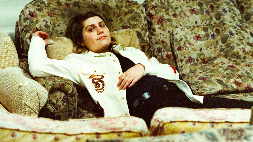 Alex Lahey is lying on a dated couch in a white denim jacket with one arm raised above her head.