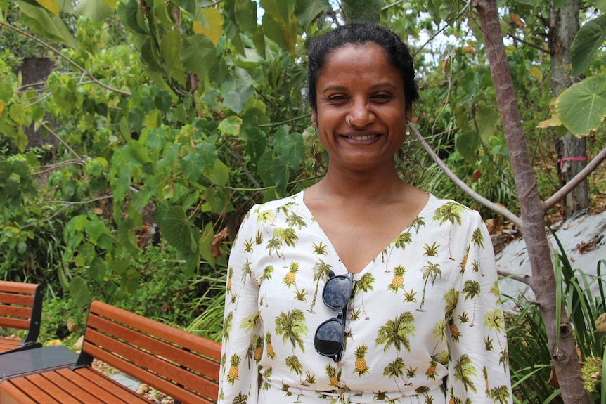 A woman in a dress with pineapples on it smiles at the camera with foliage behind her