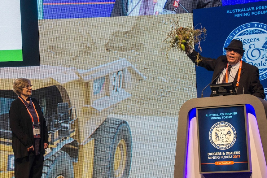 Indigenous man stands at lectern holding a bunch of native plant in his hand in the air, a woman stands to the side watching.