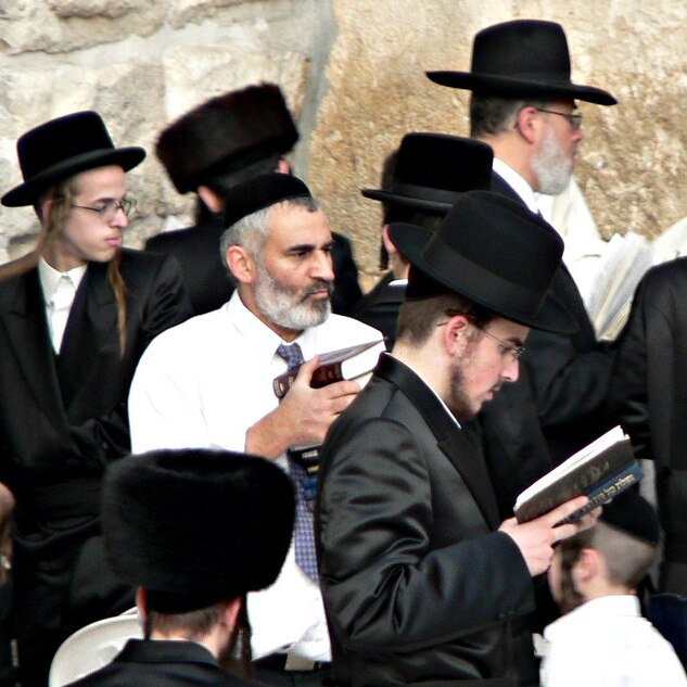 A Jewish man wearing a kippah with a white shirt is surrounded by ultra-orthodox Jews in top-hats against a wall.