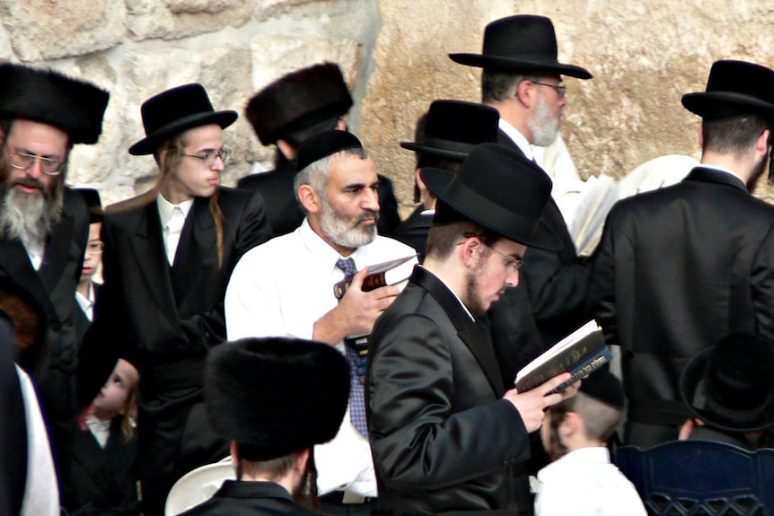 A Jewish man wearing a kippah with a white shirt is surrounded by ultra-orthodox Jews in top-hats against a wall.