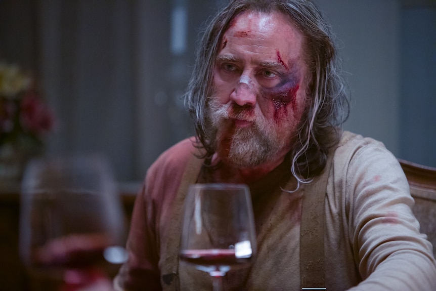 Nicolas Cage with long greying hair looks unimpressed. He's wearing a blood-soaked shirt and his face appears beaten.