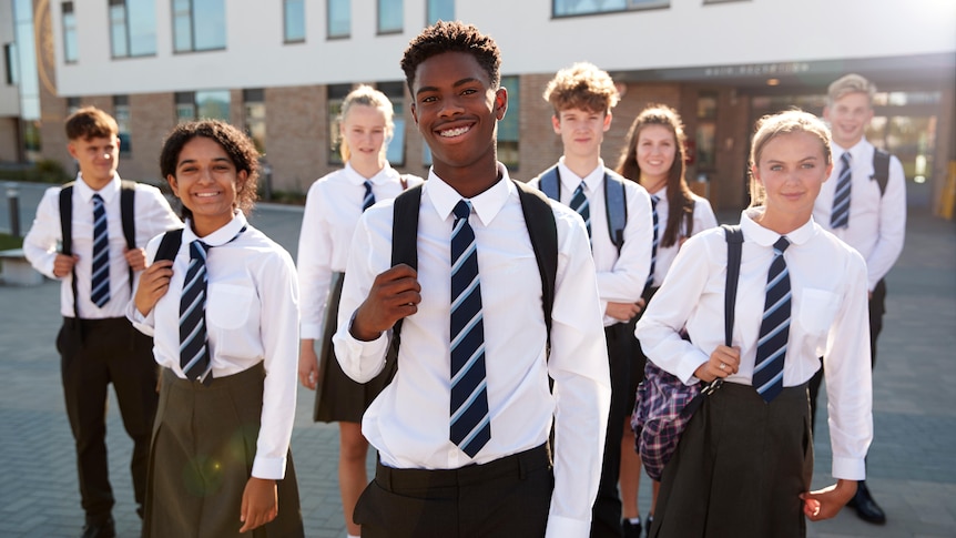 A group of smiling high school students in uniform stand in front of their school and look at the camera.