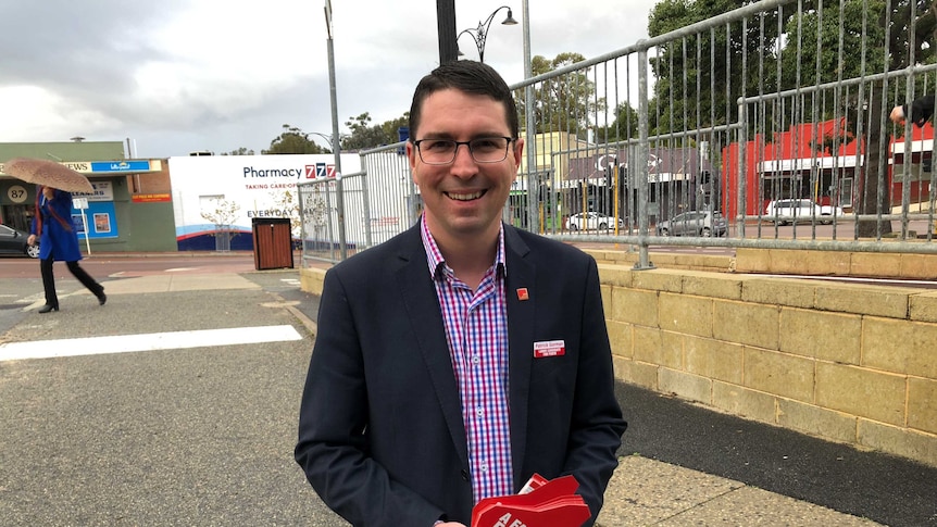 Patrick Gorman dressed in a plaid shirt and blazer smiles at the camera standing beside a fence outside