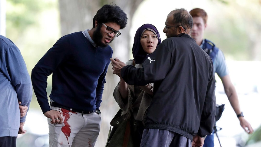 Witnesses stand around with police in the background one man has blood on his pants