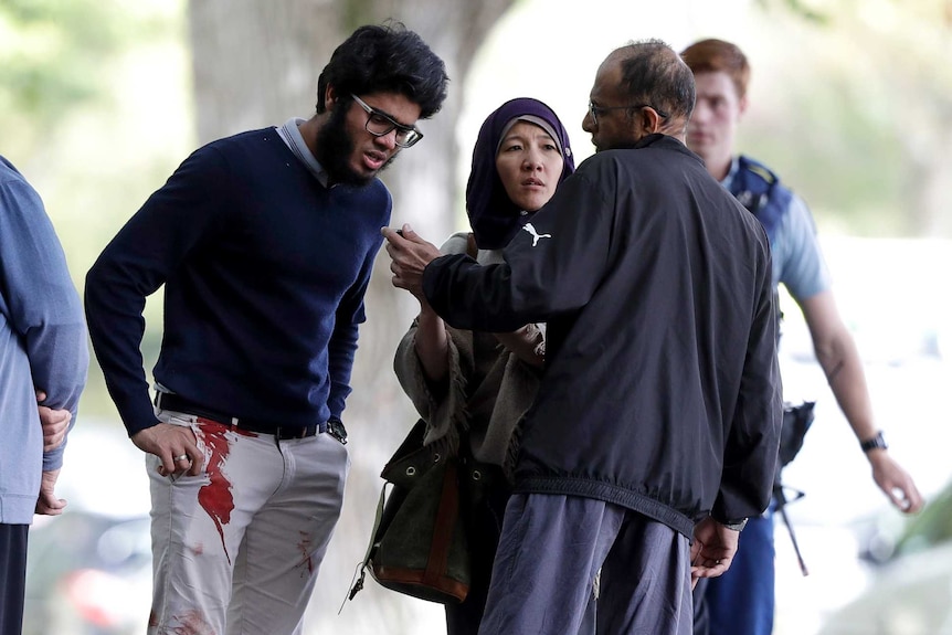 Witnesses stand around with police in the background one man has blood on his pants