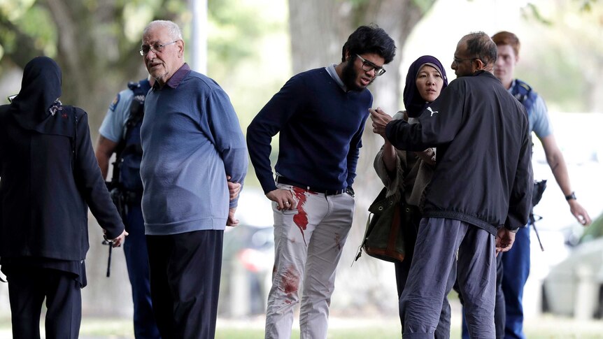 Witnesses stand around with police in the background one man has blood on his pants