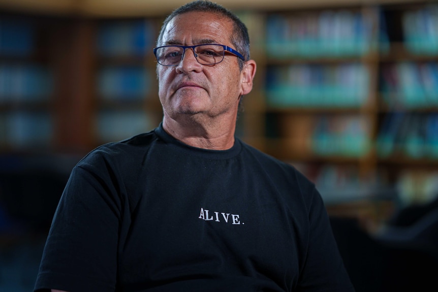 An older man wears a shirt that says "alive" on the chest area.
