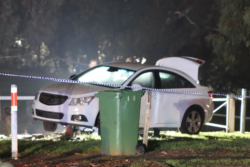 A white car on a grass verge at night with its boot up and a green bin and police tape in the foreground.
