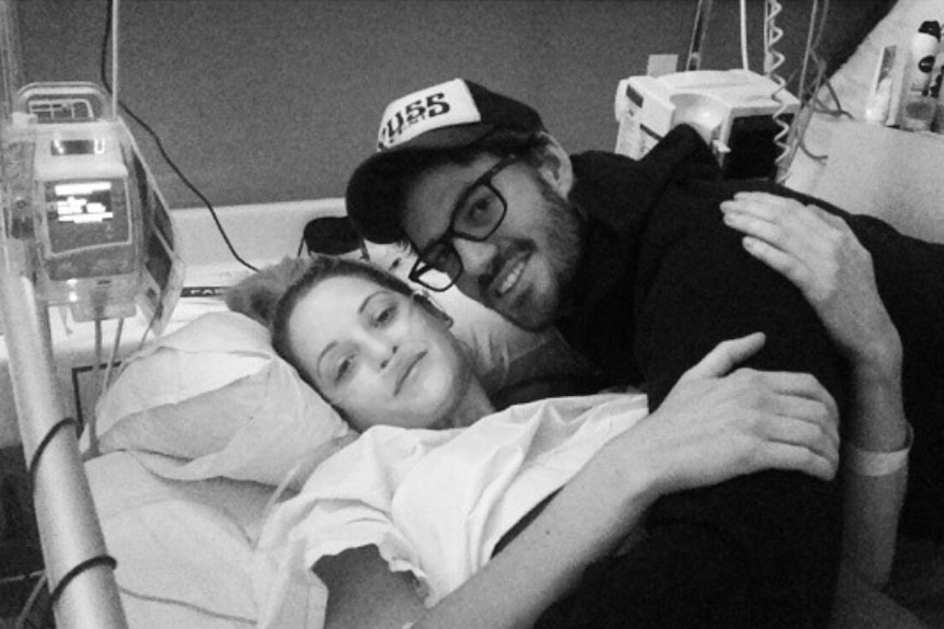 A black-and-white shot of a woman in a hospital bed being hugged by a man in a cap.