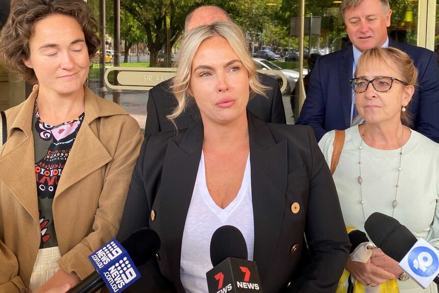 A group of men and women stand outside a court building. A woman with blonde hair looks tearful as she speaks to reporters.