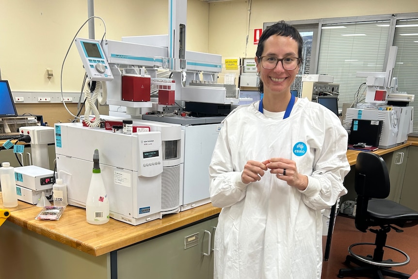 A smiling woman wearing a CSIRO-branded white lab coat and glasses standing in front of a piece of laboratory equipment