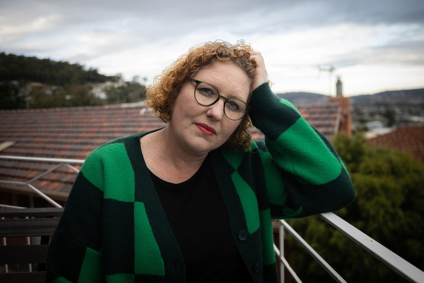 A woman with glasses and red lipstick wearing a black and green jumper leans on a balcony railing.