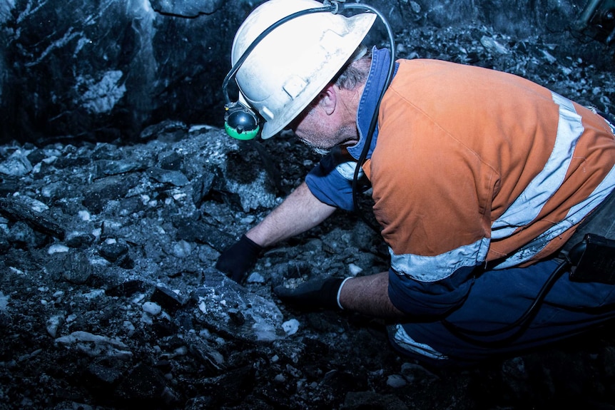 Mine working wearing hard hat and head lamp on hands and knees looking for gold-covered rocks underground.