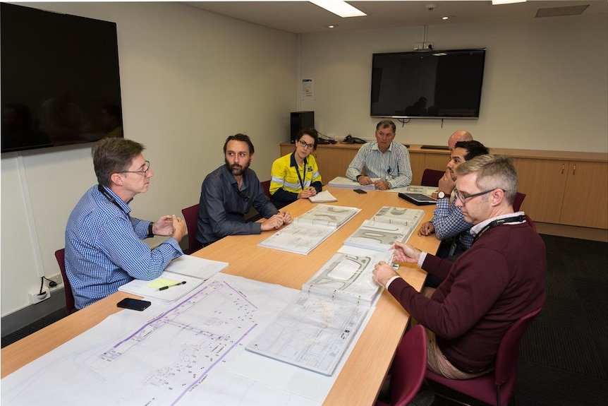 Paul Coughlan (centre) sits at a conference table during a meeting with six people, with plans on the table.