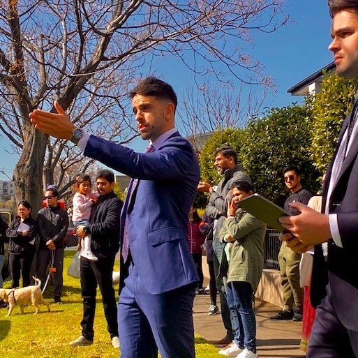 Auctioneer gestures to the crowd in front of a house he is selling. The sky is blue and a large crowd gather to watch 
